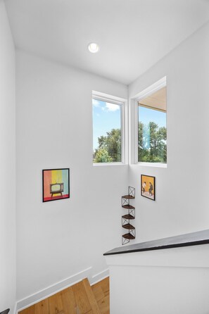 Ein Zimmer mit weißen Wänden, einer Holztreppe und zwei Fenstern mit Blick auf Grünflächen.