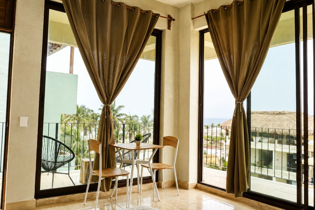 A balcony with a table and chairs, a potted plant, and a view of the sea and palm trees.