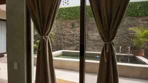 A view through a window with curtains, a stone wall, and a small pool with a plant. - CASA AMOR ZIKATELA (Colotepec)