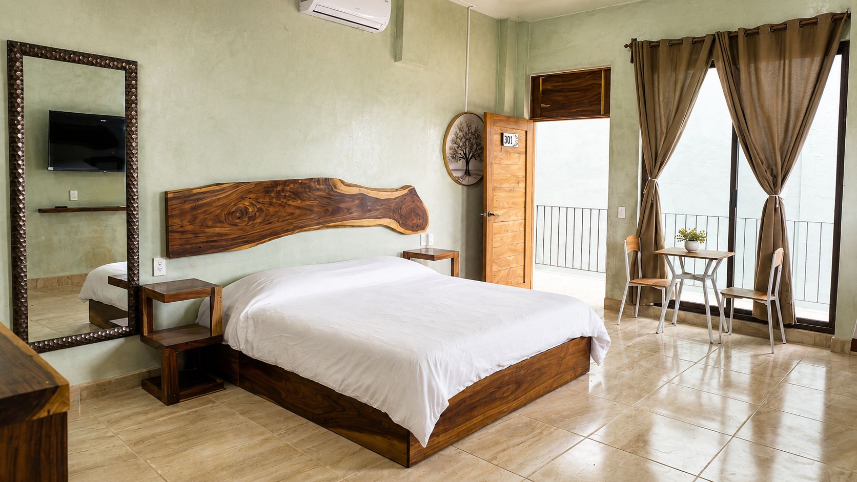 A hotel room with a wooden bed, a TV mounted on the wall, a balcony with a view, and a door with a number.