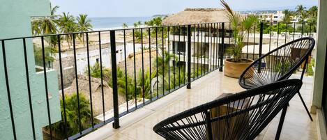 Un balcon avec deux fauteuils en osier noir, une plante en pot et une vue sur la plage et l’océan.
