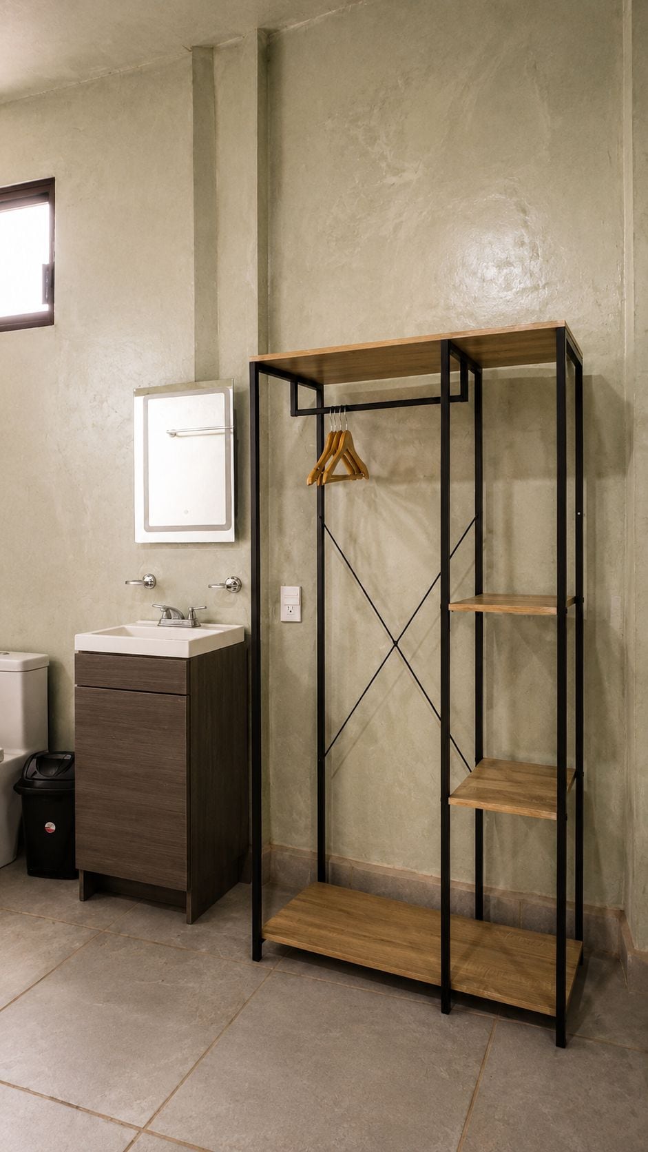 A bathroom with a wooden vanity, a white cabinet, a hanging clothes rack, and a metal shelving unit.