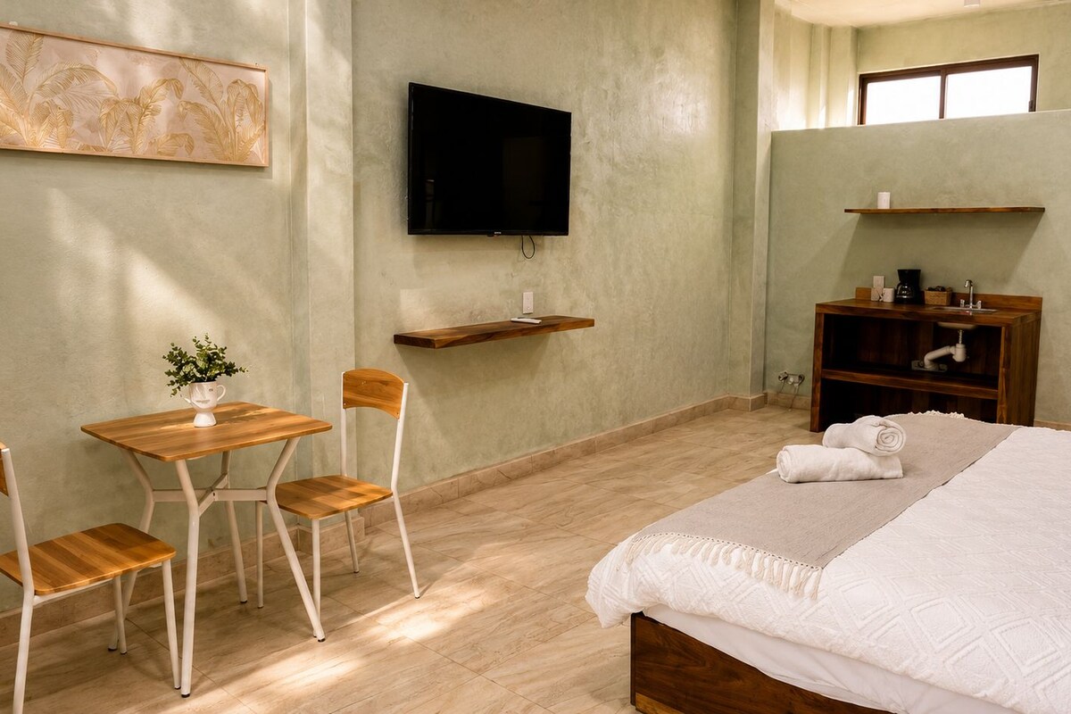A hotel room with a bed, a desk with chairs, a television, and a shelf with a coffee machine.