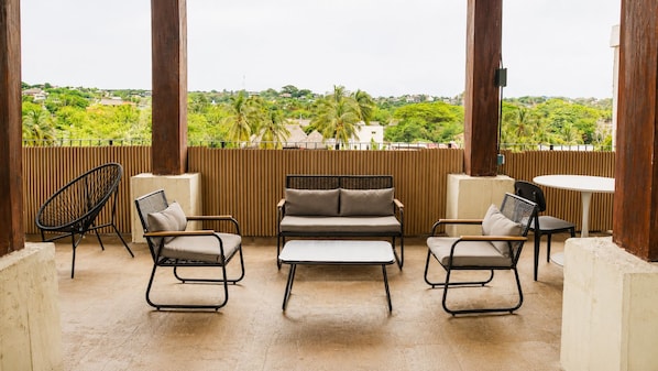 A covered outdoor seating area with a sofa, two armchairs, and a small table, overlooking a lush landscape with palm trees and buildings. - CASA AMOR ZIKATELA (Santa Maria Colotepec)