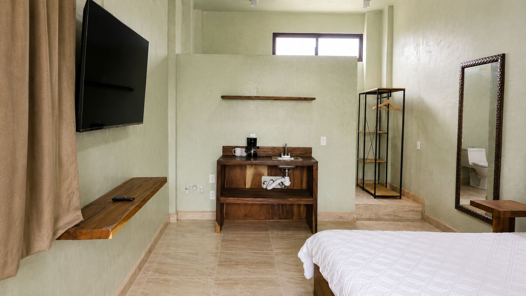 A hotel room with a bed, a wooden dresser, a TV mounted on the wall, a window, and a mirror.