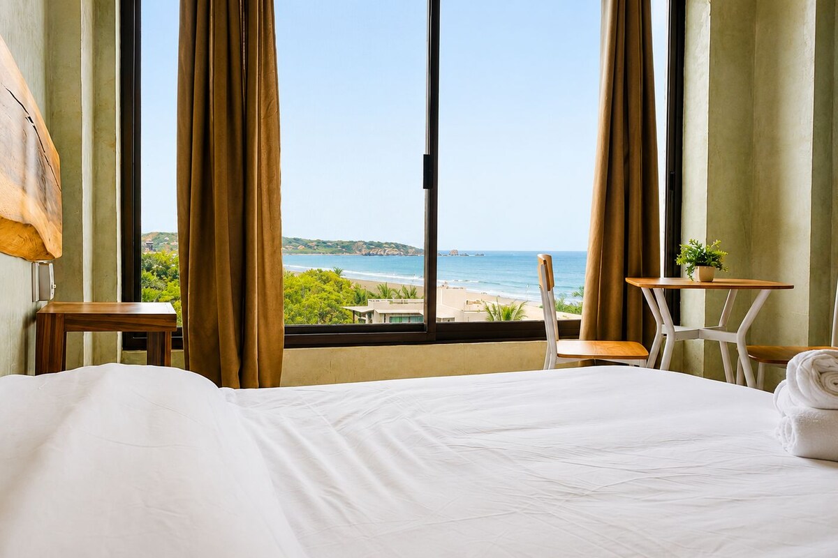 A hotel room with a large bed, a view of the beach, a small table, and a chair.