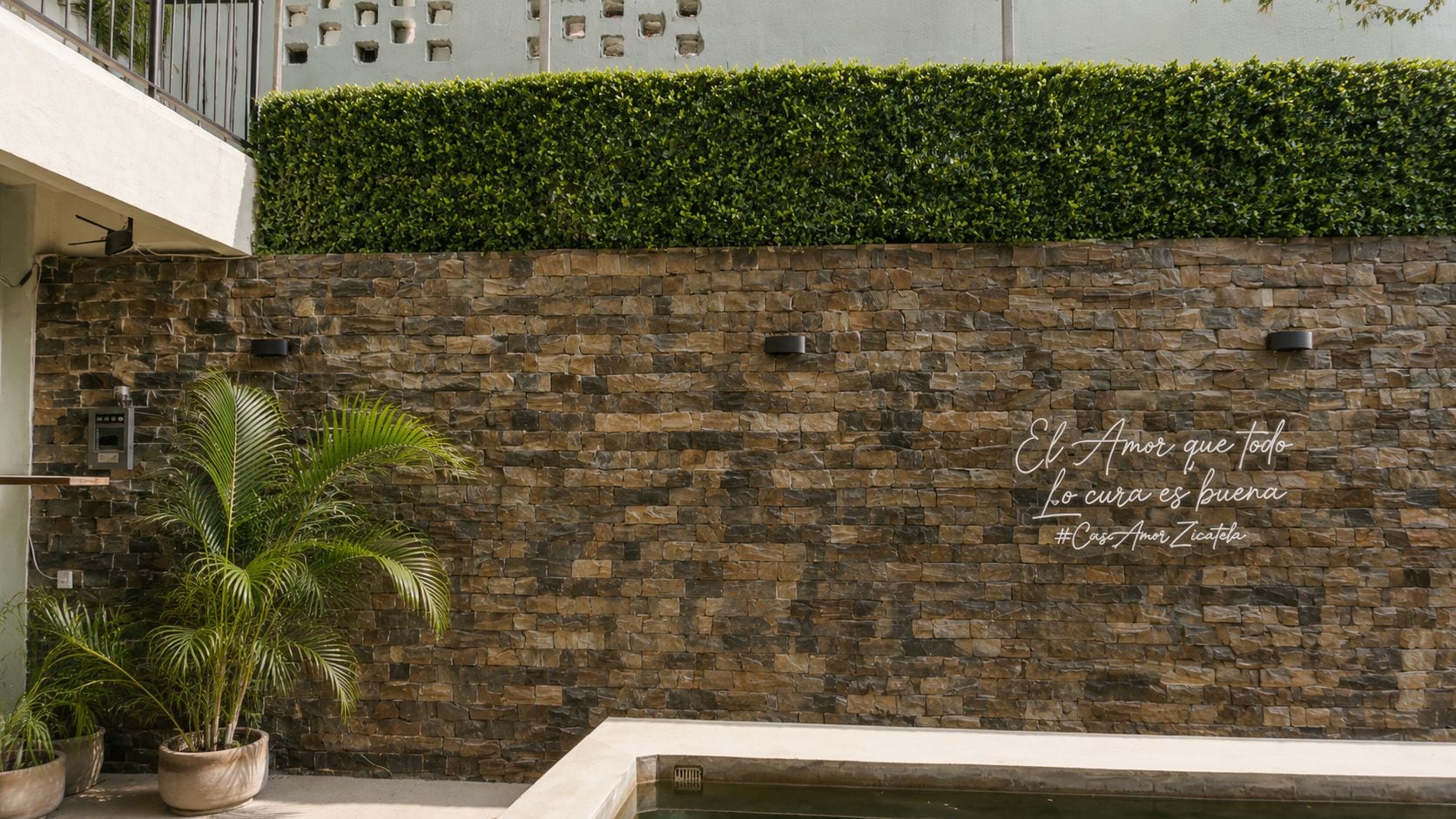 A stone wall with a quote in Spanish, a potted palm plant, and a pool.