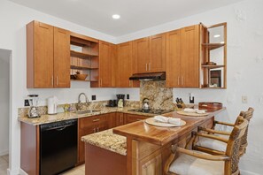 A kitchen with wooden cabinets, granite countertops, a built-in dishwasher, and a bar area with a chair. - Kapalua 14T3 | Spacious 3BR Maui Vacation Home (Lahaina)