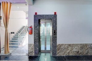 Entrada de hotel con piso de mármol, puerta de ascensor de vidrio y extintor colgado en la pared.