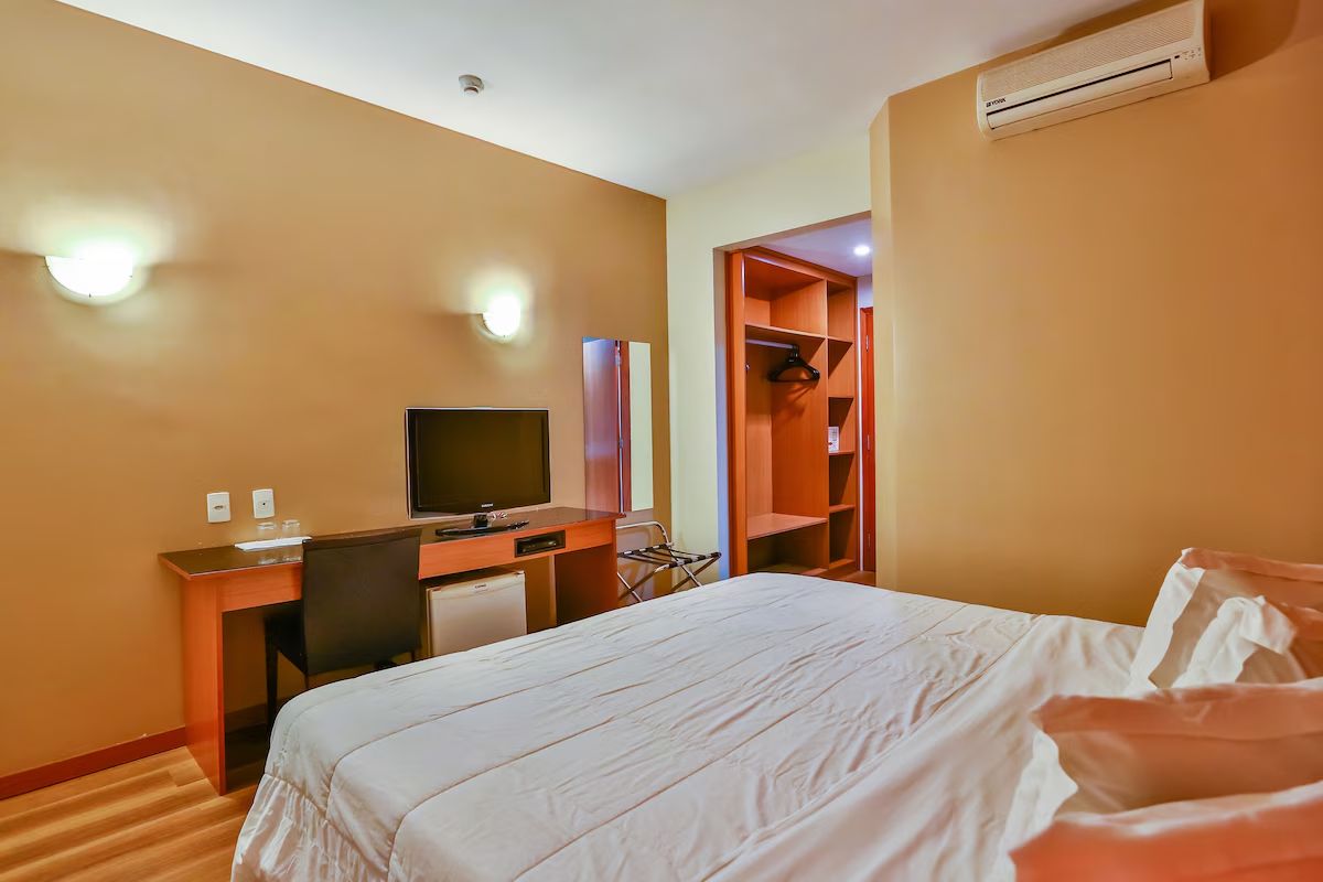 Quarto de hotel com cama, escrivaninha com computador, televisão e guarda-roupa.