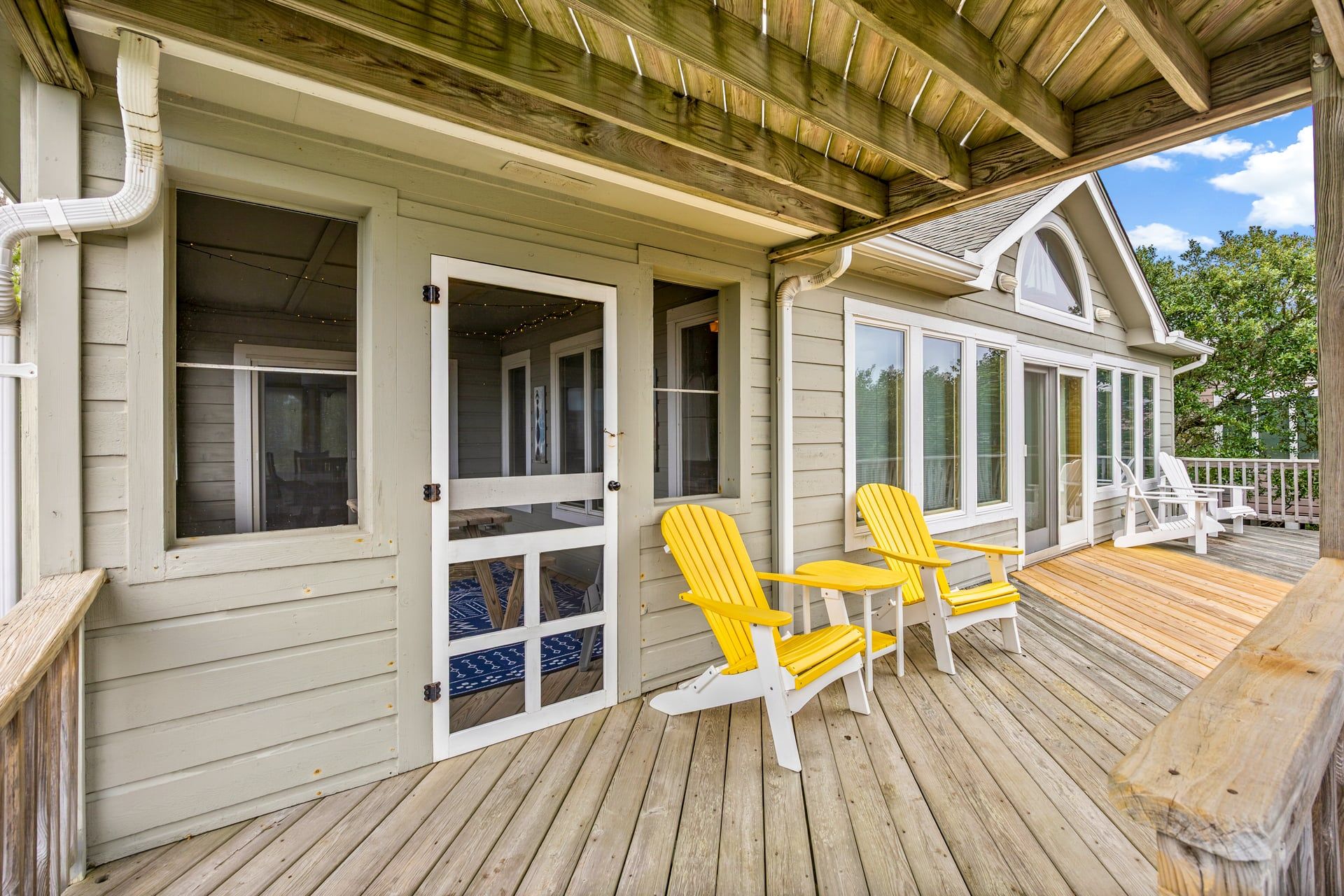 A wooden deck with two yellow Adirondack chairs, a covered porch, and a large window overlooking a green landscape.