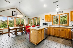 A kitchen with wooden cabinets, a central island, a dining area with chairs, and large windows with a view of greenery. - Turtle's Cove Corolla retreat with beach access, hot tub, and views. (Corolla)