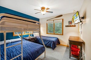 A room with bunk beds, a ceiling fan, a window with a view of trees, a wooden table, and a wall-mounted TV. - Turtle's Cove Corolla retreat with beach access, hot tub, and views. (Corolla)
