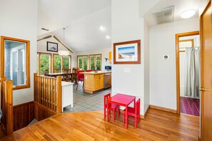 A spacious room with a wooden floor, a dining area with red chairs, a kitchen with wooden cabinets, and a view of greenery through large windows. - Turtle's Cove Corolla retreat with beach access, hot tub, and views. (Corolla)