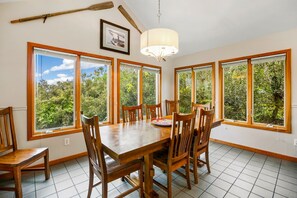 Un comedor con mesa y sillas de madera, amplias ventanas con vista a follaje, una araña y un cuadro en la pared.