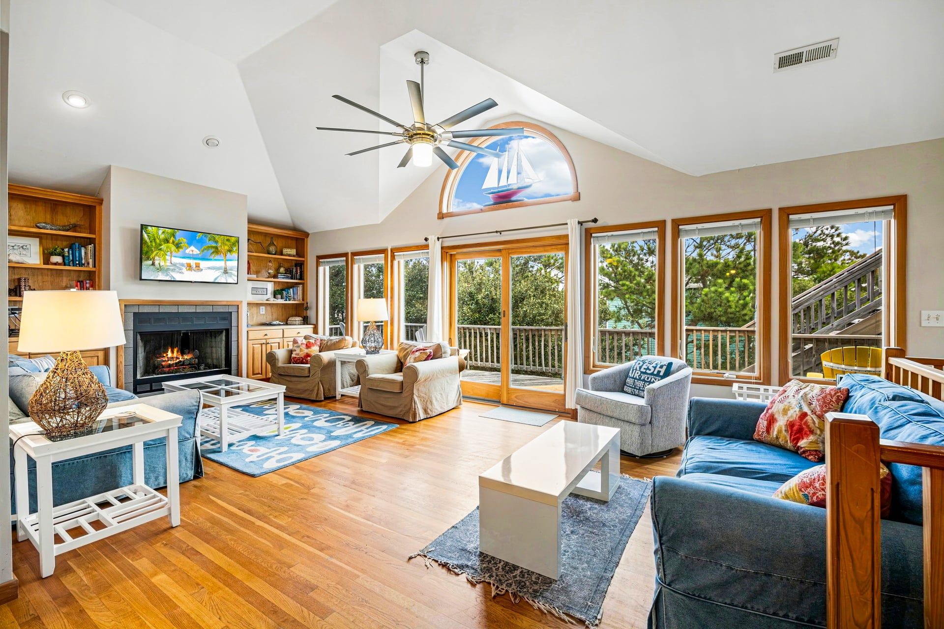 A spacious living room with a fireplace, comfortable seating, a ceiling fan, and large windows offering views of the outdoors.