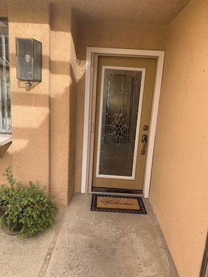 A beige textured wall, a glass door with a metal handle, a welcome mat, and a small potted plant. - Our Little Gem In The Desert (Rancho Mirage)
