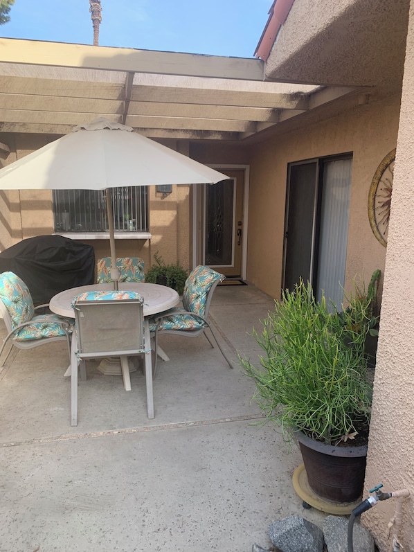 A patio with a table and chairs, a covered umbrella, a potted plant, and a sliding glass door. - Our Little Gem In The Desert (Rancho Mirage)