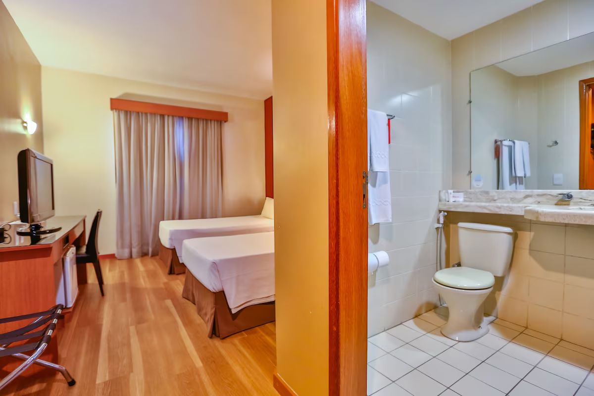 Ein Hotelzimmer mit einem Holztisch, einem Fernseher, zwei Betten, einem Badezimmer mit Toilette und Waschbecken sowie einem großen Spiegel.