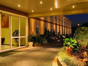 Entrada de hotel con un camino empedrado, plantas en macetas y un edificio con ventanas amplias y un balcón.
