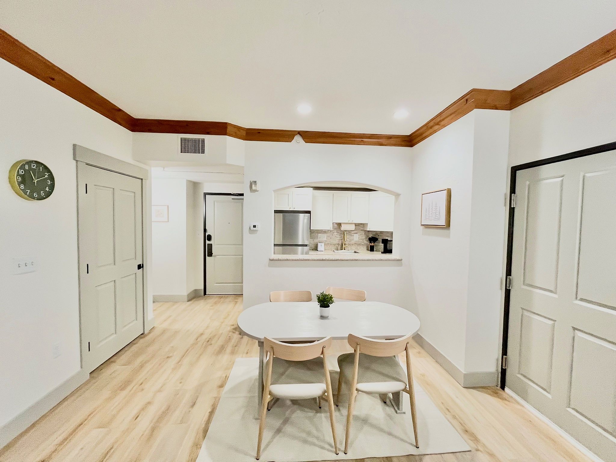 Un coin repas avec une table ronde et quatre chaises, une cuisine avec des meubles blancs et une horloge au mur.