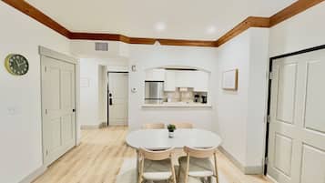 Un coin repas avec une table ronde et quatre chaises, une cuisine avec des meubles blancs et une horloge au mur.