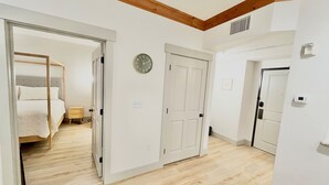 A hotel room with a wooden floor, a bed, a bedside table, a clock, and a door leading to another room.