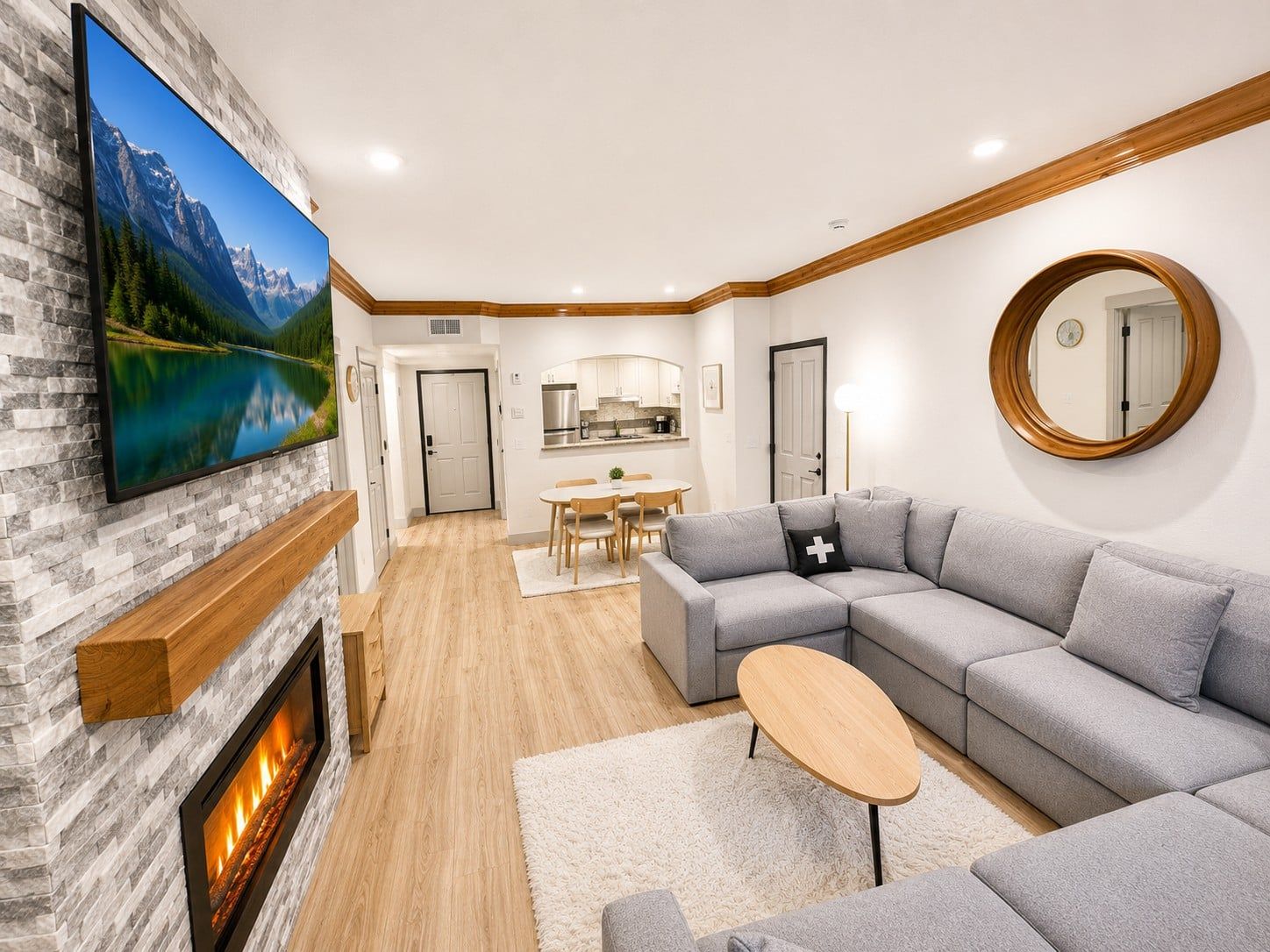 Una sala de estar moderna con chimenea, un televisor de pantalla plana grande montado en una pared de piedra, un sofá seccional gris, un espejo redondo y un comedor con una mesa y sillas de madera.