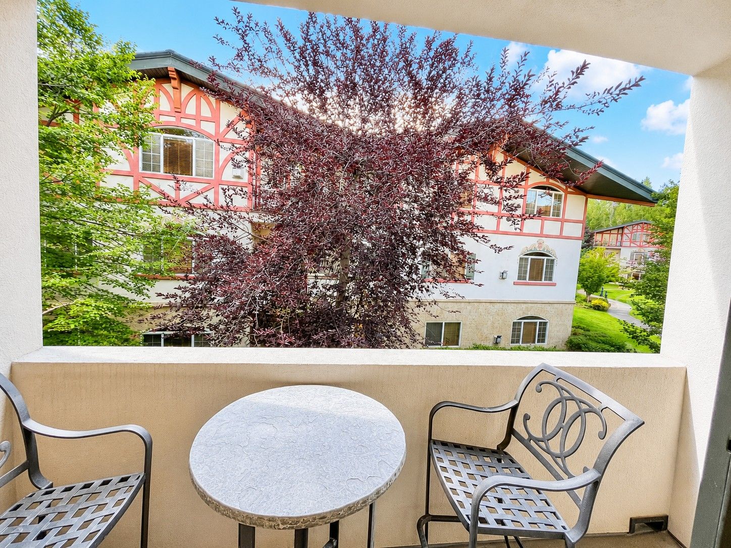Un balcon avec une table et deux chaises, donnant sur un immeuble à la façade rouge et blanche et sur un arbre aux feuilles violettes.