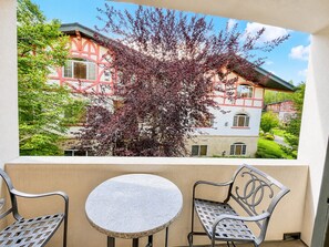 Un balcone con un tavolo e due sedie, affaccia su un edificio con facciata rossa e bianca e un albero dalle foglie viola.
