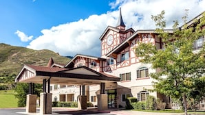 A traditional chalet-style hotel with a prominent clock tower, a covered entrance, and a mountainous backdrop.