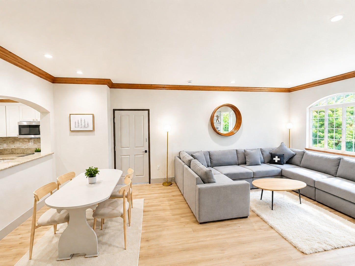 Un salon moderne avec un canapé d’angle gris, une table basse ronde et un coin repas comprenant une table et des chaises blanches.