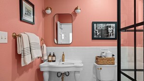 Un baño con pared rosa, baldosas blancas, lavamanos con detalles dorados, un inodoro y un cuadro enmarcado sobre el espejo.