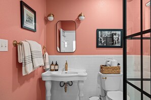 A bathroom with a pink wall, white tiles, a sink with gold fixtures, a toilet, and a framed picture above the mirror.