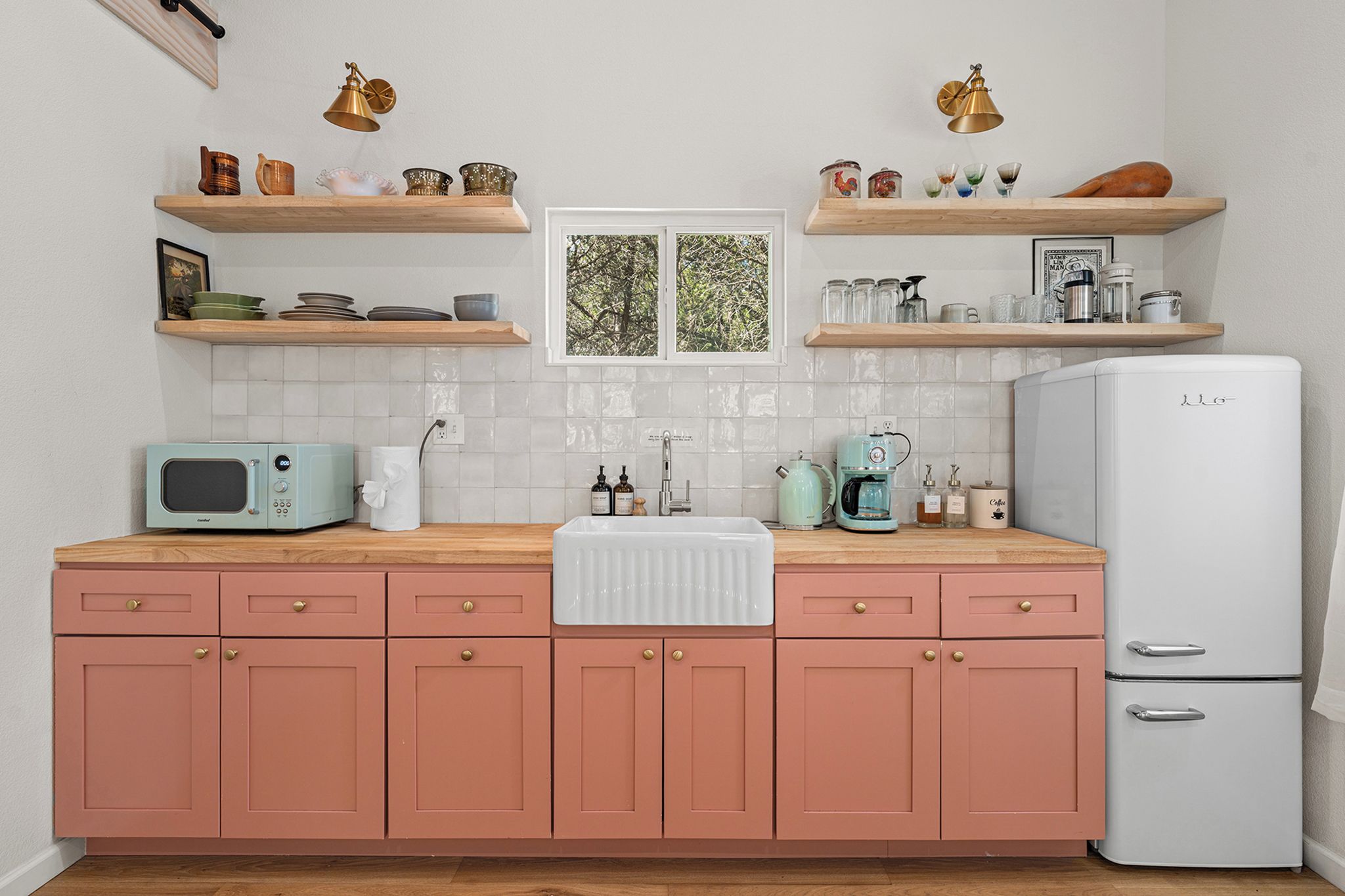 Una cocina con armarios rosas, un refrigerador blanco, un microondas, un lavamanos y varios utensilios de cocina en estantes.