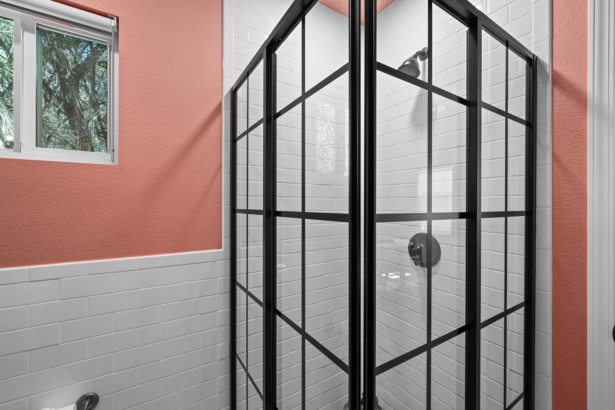 A bathroom with a glass-enclosed shower, white tiled walls, and a window with a view of trees.
