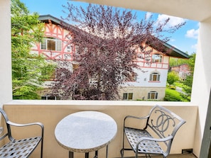 Un balcone con un tavolo e due sedie, affaccia su un edificio con facciata rossa e bianca e un albero dalle foglie viola.