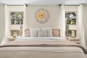 A bedroom with a large bed, two bedside tables, a wall-mounted artwork, and a view through the windows. - Gathering Oak | Cozy Fire Pit & Fall Getaway (Wimberly)