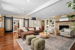 A spacious living room with a brown leather sofa, a round wooden coffee table, and a grey armchair. A large rug covers the floor, and a fireplace is visible in the background. - Gathering Oak | Cozy Fire Pit & Fall Getaway (Wimberly)
