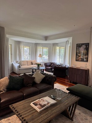 A spacious living room with a brown leather sofa, a white armchair, and a dark brown leather armchair. There is a wooden coffee table with a magazine and a remote control. - Harrowby Country House. New Magnesium Pool. Outdoor firepit. Sleeps 11. (Broke)