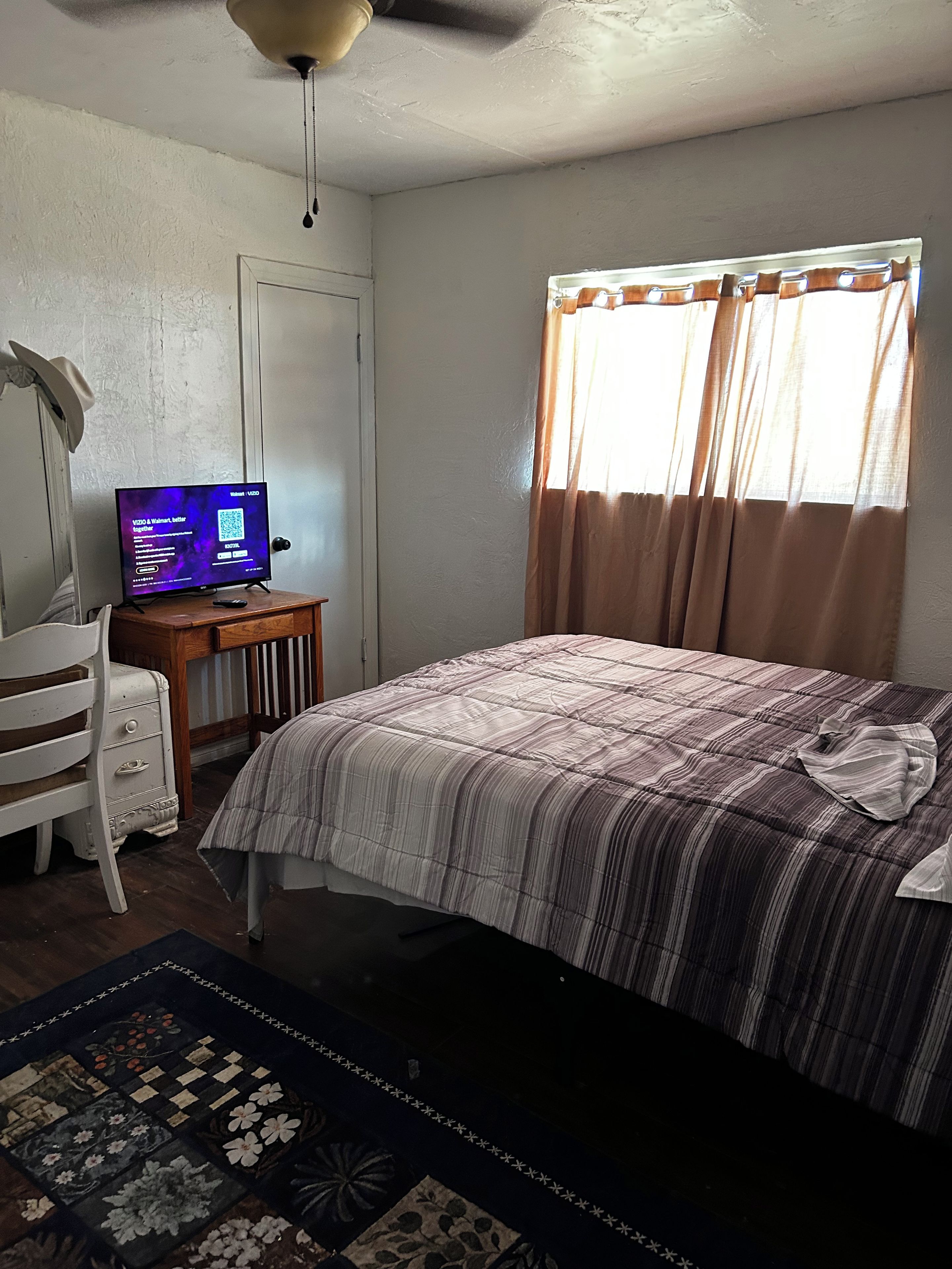 A bedroom with a bed, a desk with a computer, a chair, and a window with curtains.