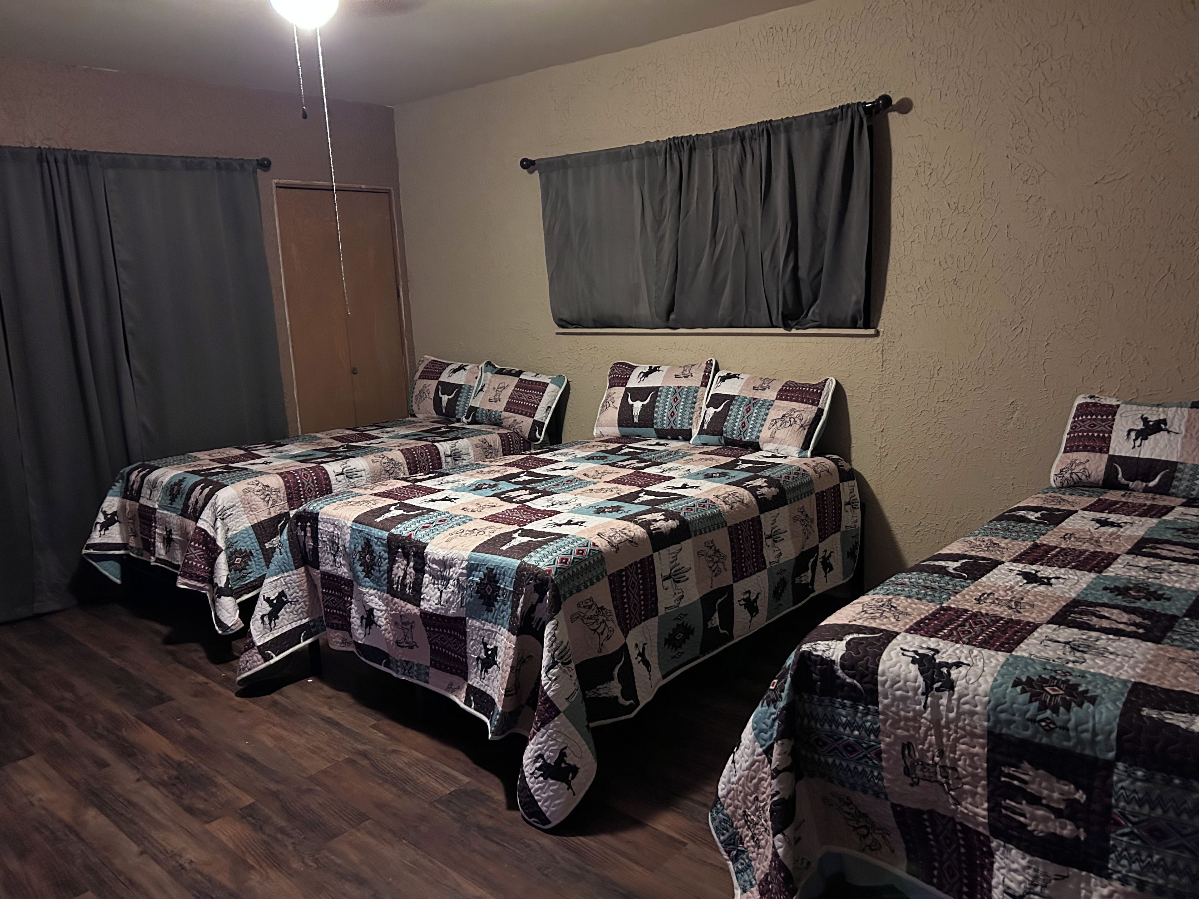 A room with two beds, each with a patterned quilt, a wooden floor, a door, and a window with curtains.