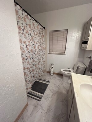 Une salle de bain comprenant des toilettes, un bidet, un rideau de douche, une fenêtre avec des stores et un sol en marbre.