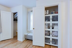 A room with a wooden floor, a white door, a closet with hanging clothes, and a bed with a white mattress.
