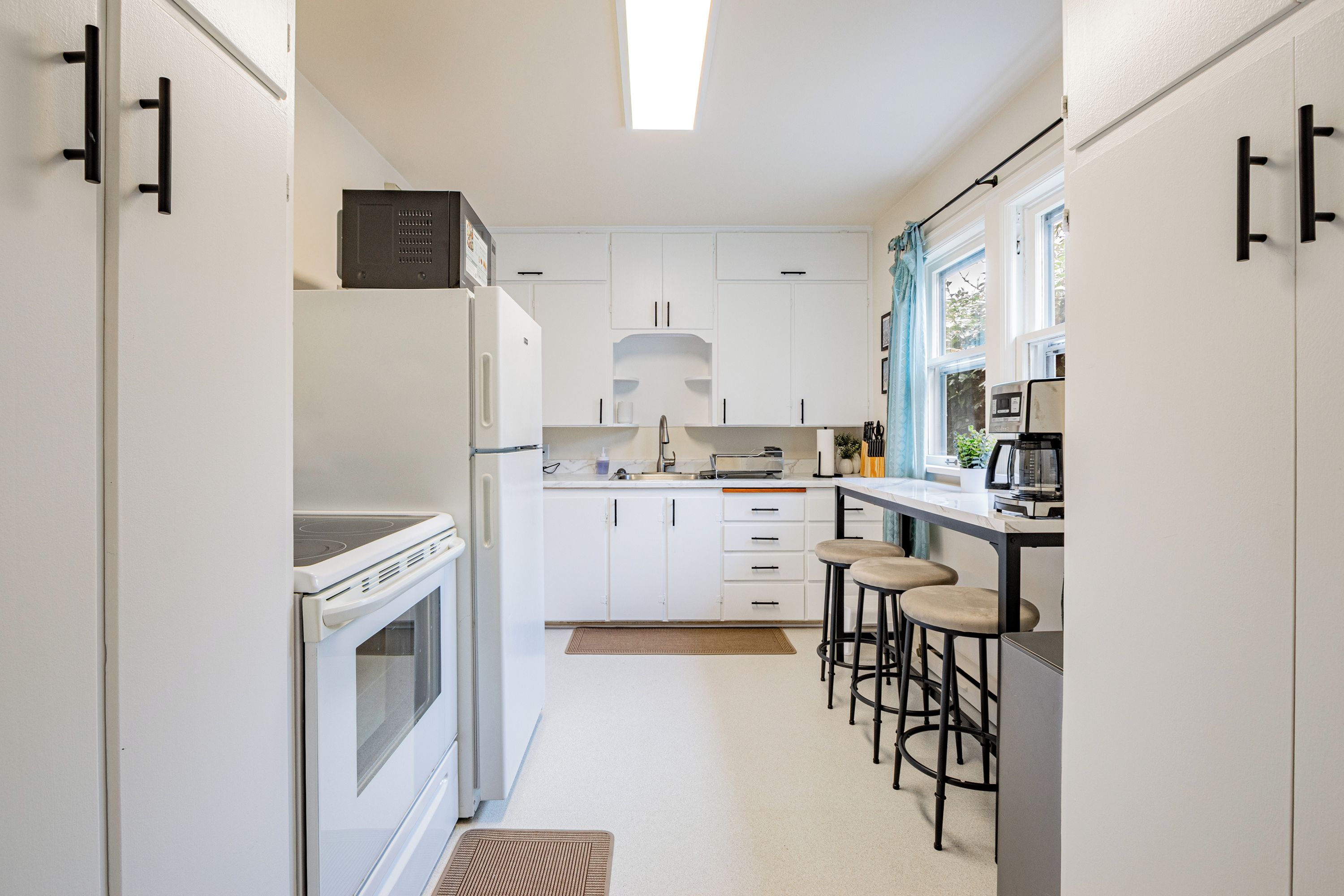 A compact kitchen with white cabinetry, a built-in oven, a microwave, and a coffee machine. There are bar stools and a window with curtains.