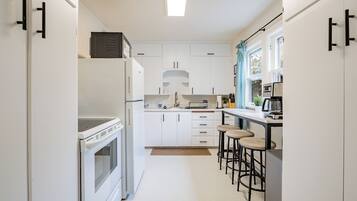 A compact kitchen with white cabinetry, a built-in oven, a microwave, and a coffee machine. There are bar stools and a window with curtains.