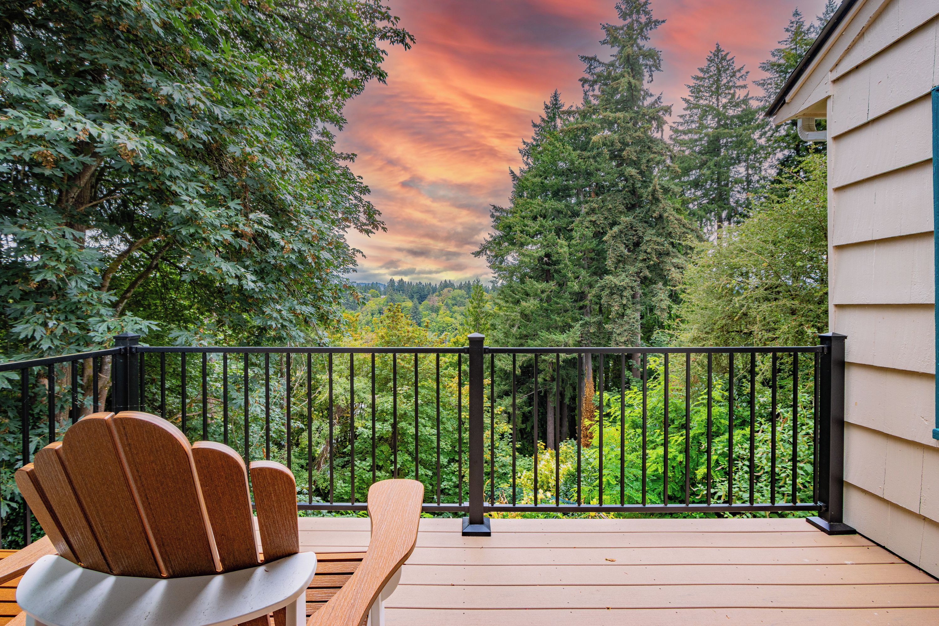 Una terraza de madera con barandilla negra, una silla Adirondack de madera y vistas a un paisaje boscoso bajo un cielo colorido.