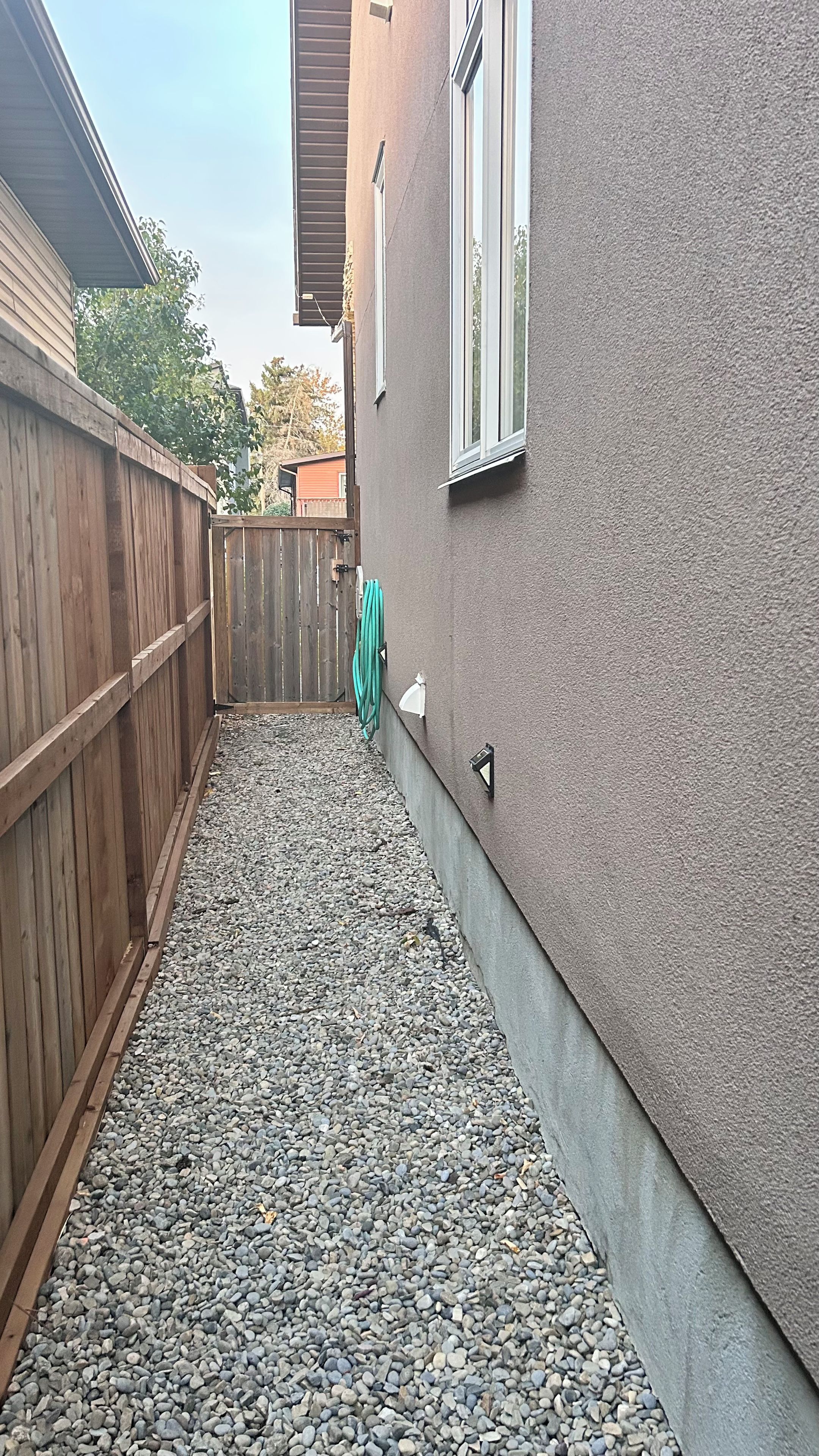 Um espaço exterior estreito com cerca de madeira, chão de cascalho e uma casa com janela e luminária fixada na parede.