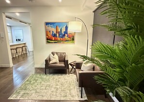 Ein Wohnzimmer mit Stadtbildgemälde, einer Couch, einem Sessel, einem Couchtisch und einer großen Zimmerpflanze.
