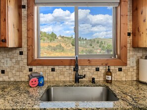 A kitchen with a granite countertop, stainless steel sink, and a window with a view of a hilly landscape.